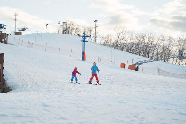 Безликий родитель в теплой спортивной одежде и шлеме учит маленького ребенка кататься на лыжах вдоль снежного склона на зимнем горнолыжном курорте — стоковое фото