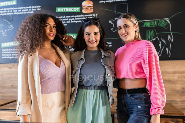 Jovens amigas multirraciais felizes em roupas elegantes que estão perto da placa de menu no restaurante moderno e olhando para a câmera — Fotografia de Stock