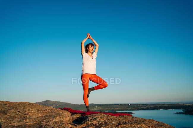 Joven viajera en ropa deportiva de pie en pose de árbol con los brazos arriba asana mientras practica yoga en la montaña rocosa sobre la costa contra el cielo azul sin nubes - foto de stock