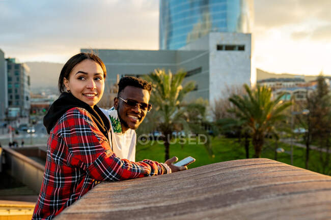Happy multiracial couple in stylish apparel with cellphone speaking in town under cloudy sky in twilight — Stock Photo