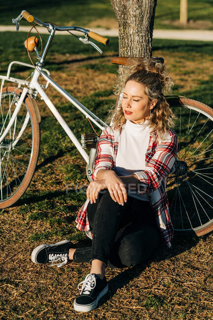 Alto ângulo de mulher sonhadora sentada perto de bicicleta perto da árvore no parque e desfrutando de tempo ensolarado com olhos fechados — Fotografia de Stock