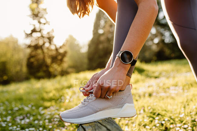 Vista lateral de una deportista anónima en un rastreador portátil atando cordones de zapatos en el calzado antes de hacer ejercicio en el prado a la luz del sol - foto de stock