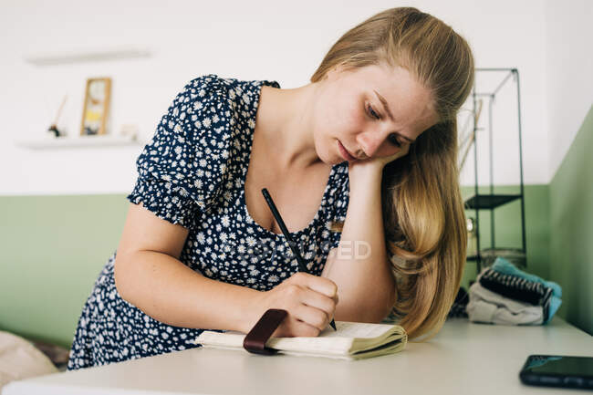 Молода жінка спирається рукою на столі, роблячи нотатки в блокноті вдома в денний час — стокове фото