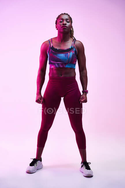 Giovane atleta donna afro-americana muscolosa in abiti sportivi con trecce afro e mani sui fianchi distogliendo lo sguardo nella luce al neon — Foto stock