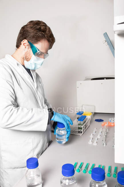 Seitenansicht eines männlichen Biologen in Schutzbrille und Maske, der eine Flasche mit Flüssigkeit am Tisch im Cannabis-Labor öffnet — Stockfoto