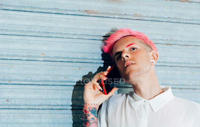Transsexual man in trendy wear with tattoo and pink hair speaking on cellphone while looking away — Stock Photo