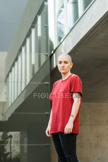 Junge, emotionslose lesbische Frau im roten T-Shirt blickt gegen Hausfassade in der Stadt in die Kamera — Stockfoto