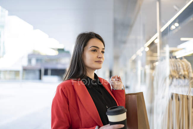 Весёлая женщина-покупатель с бумажными пакетами для покупок и кофе на вынос стоит рядом с витриной магазина в городе — стоковое фото
