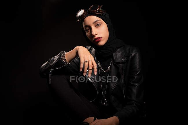 Confident young Muslim female in leather jacket headscarf and creative sunglasses sitting against black background in studio — Stock Photo