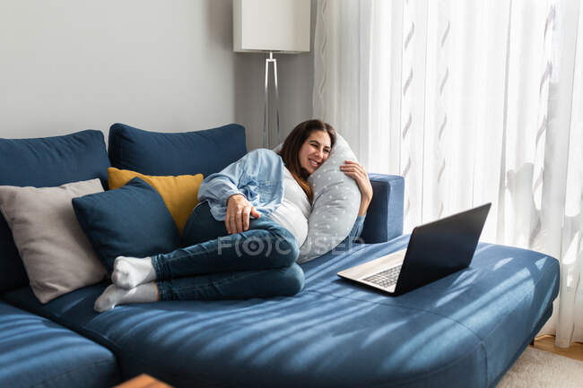 Entzückte schwangere Frau liegt auf bequemem Sofa und schaut lustige Videos auf Netbook, während sie zu Hause entspannt — Stockfoto