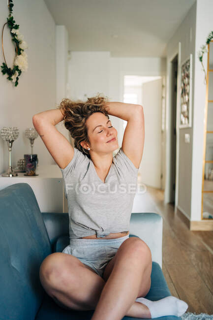 Positive attractive female in shorts sitting on cozy couch in living room stretching with arms up with eyes closed — Foto stock