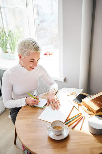 Astróloga sonriente tomando notas en bloc de notas en el escritorio con una taza de café en casa a la luz del sol - foto de stock