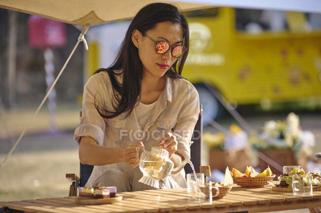 Schöne ethnisch-asiatische Frau sitzt am Tisch und serviert Tee auf Glaskanne, während sie eine entspannte Zeit auf dem Campingplatz während der Ferien — Stockfoto