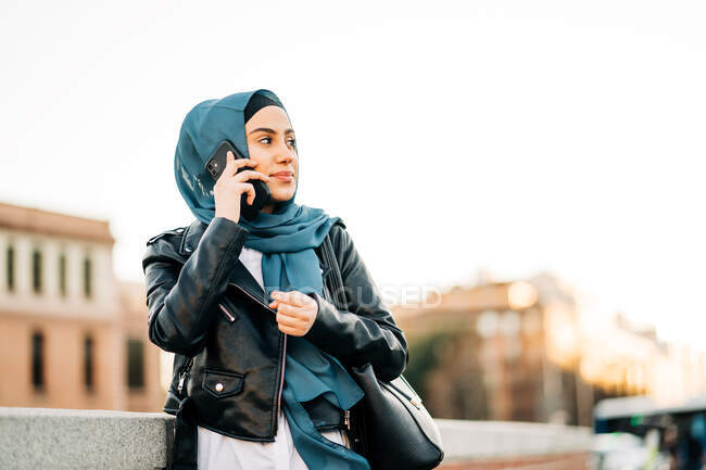 Mujer musulmana alegre en pañuelo tradicional de pie en la calle de la ciudad y hablando por teléfono móvil mientras mira hacia otro lado - foto de stock