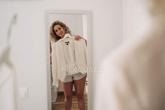 Schnurgerade junge stylische Frau mit lockigem Haar steht neben Spiegel und probiert trendigen Blazer bei Tageslicht zu Hause an — Stockfoto