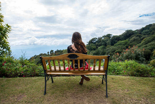 Back view of anonymous female tourist sitting with crossed legs on bench against lush trees under cloudy sky in Thailand — Stock Photo