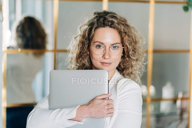 Самозабезпечена молода жінка-підприємець з кучерявим світлим волоссям у білій блузці та окулярах, дивлячись на камеру, стоячи в сучасному робочому просторі з ноутбуком в руках — стокове фото