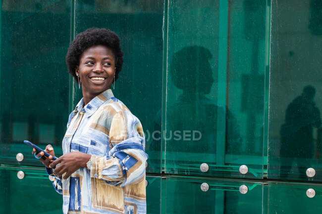 Веселая молодая афроамериканка с прической, одетая в стильную красочную рубашку, стоящую у стеклянной стены и просматривающую мобильный телефон, отворачивающуюся — стоковое фото