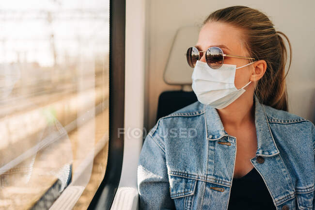 Mujer joven en chaqueta de mezclilla y máscara facial protectora mirando hacia otro lado en la ventana del tren mientras viaja - foto de stock