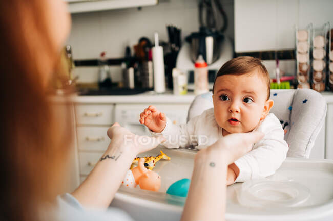 Cute baby in white shirt sitting in baby feeding chair in modern kitchen — Stock Photo