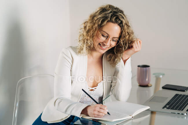 Young female freelancer with curly blond hair in casual clothes and eyeglasses taking notes in planner and looking away while working remotely using laptop at home — Stock Photo