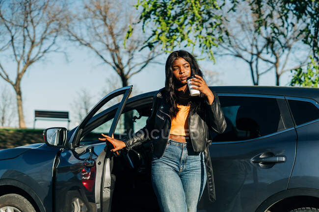 Уверенная афроамериканка с выпивкой на вынос, стоящая возле черного роскошного автомобиля и отводящая взгляд — стоковое фото