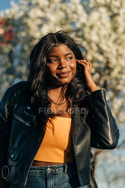 Hermosa mujer afroamericana de pie en el floreciente parque de primavera y disfrutando del clima soleado mirando a la cámara - foto de stock