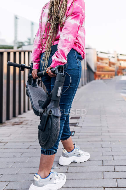 Анонимная женщина на урожае в повседневной одежде стоит с современным скутером на мощеной дорожке в городе — стоковое фото