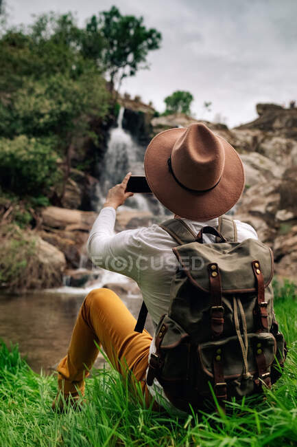 Обратно вид безликого мужчины путешественника в повседневной одежде и шляпе, фотографирующего на смартфон быстрый каскад, падающий с скалистых образований в зеленой природе — стоковое фото