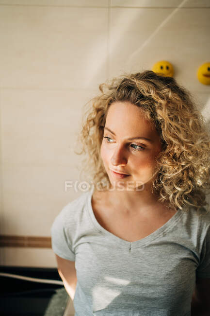 Verträumtes junges Weibchen mit lockigem Haar steht zu Hause vor hell gekachelter Wand und schaut in angenehmen Gedanken weg — Stockfoto