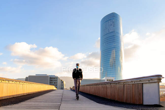 Empreendedor masculino étnico irreconhecível em máscara estéril montando scooter elétrico na passarela da ponte da cidade contra edifícios sob céu azul nublado durante a pandemia — Fotografia de Stock