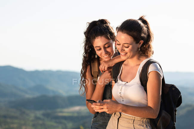 Felice giovani viaggiatori femminili in abiti estivi utilizzando smartphone insieme mentre in piedi su un lussureggiante terreno collinare soleggiato — Foto stock
