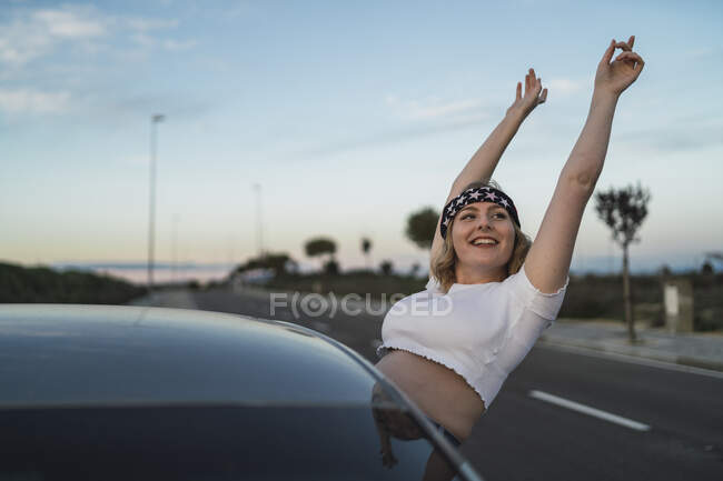 Jeune femme en tenue décontractée et bandeau imprimé drapeau américain penché par la fenêtre de la voiture et levant les mains tout en profitant de la liberté pendant le voyage sur la route au coucher du soleil — Photo de stock