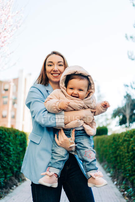 Jovem mãe alegre em roupas casuais olhando para a câmera carregando adorável bebê alegre enquanto está de pé na calçada da cidade no dia ensolarado da primavera — Fotografia de Stock