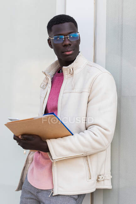 Stylish African American male entrepreneur reading documents on clipboard while standing on background of contemporary office building — Stock Photo