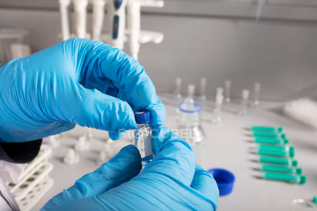 Анонимный ученый в одноразовых перчатках с жидкостью в маленькой прозрачной бутылке работает в лаборатории — стоковое фото