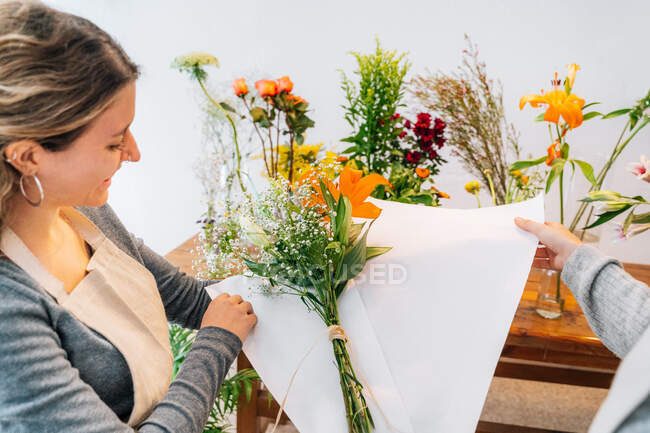 Vista laterale di coltura fiorista femminile felice in grembiule avvolgente bouquet di giglio d'arancia fresca e gypsophila in carta con collega anonimo — Foto stock