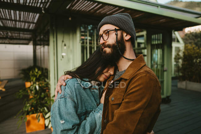 Vista lateral de la pareja joven y elegante abrazándose suavemente mientras está de pie en la terraza de madera cerca de la casa - foto de stock