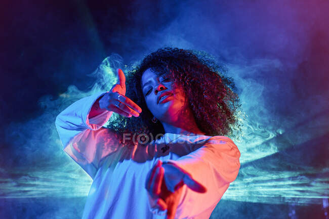 Портрет энергичной молодой афроамериканской женщины в белом, вытянувшей руки к камере во время танцев в темной студии в неоновом свете — стоковое фото