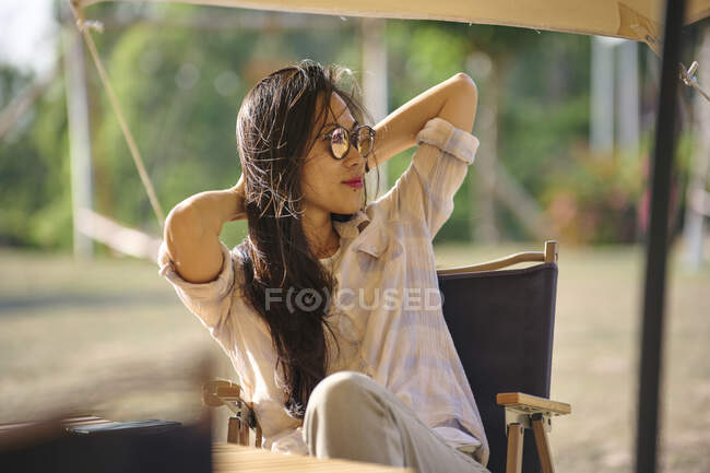 Belle femme asiatique ethnique dans des lunettes de soleil assis à la table tout en ayant un moment de détente dans la zone de camping pendant les vacances en regardant loin — Photo de stock
