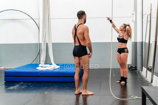 Full length giovane donna sportiva in pantaloncini sportivi indicando lontano mentre in piedi vicino istruttore maschile nel moderno centro fitness leggero — Foto stock