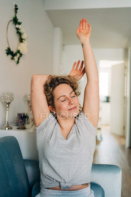 Mujer atractiva positiva en pantalones cortos sentados en un acogedor sofá en la sala de estar que se extiende con los brazos hacia arriba con los ojos cerrados - foto de stock