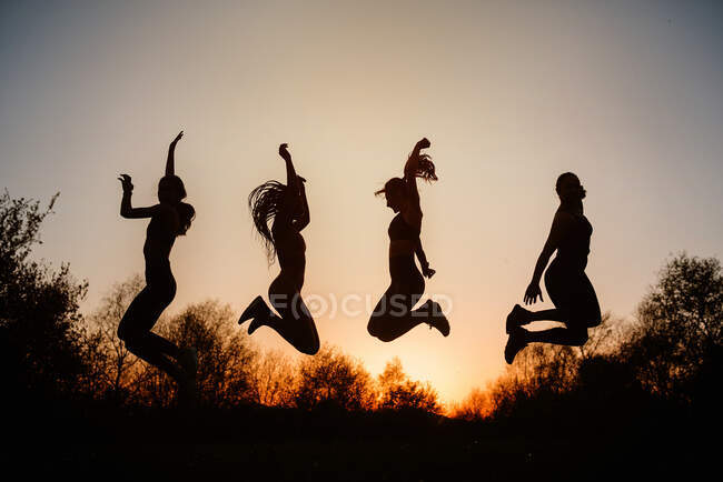 Siluetas de hembras saltando sobre el suelo contra el cielo al atardecer en el parque - foto de stock