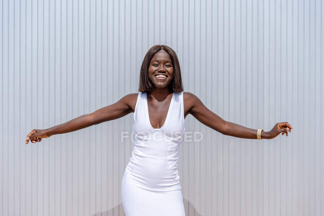 Выразительная красивая афроамериканка в модном белом платье, вытянув руки, смотрит в камеру с красивой улыбкой, стоя напротив стены здания на солнечной улице. — стоковое фото