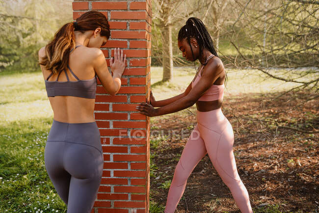 Side view of young multiethnic sportswomen in activewear working out between rough post while looking down in city park — Stock Photo