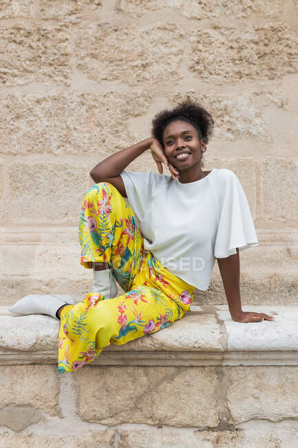 Smiling African American female in trendy outfit sitting against wall and looking away in sunny summer — Stock Photo