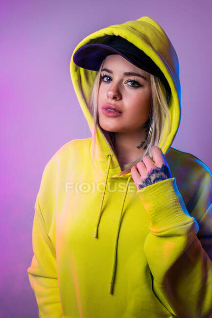 Unbewegte Frau in Streetstyle-Kapuzenpulli und Mütze blickt in die Kamera auf lila Hintergrund im Studio mit Neon-Beleuchtung — Stockfoto