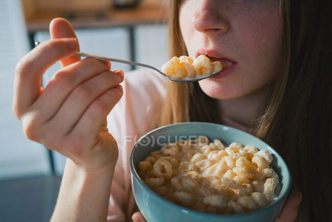 Обрізати молоду жінку з ложкою і мискою насолоджуючись смачними кільцями кукурудзи, дивлячись на камеру на кухні — стокове фото