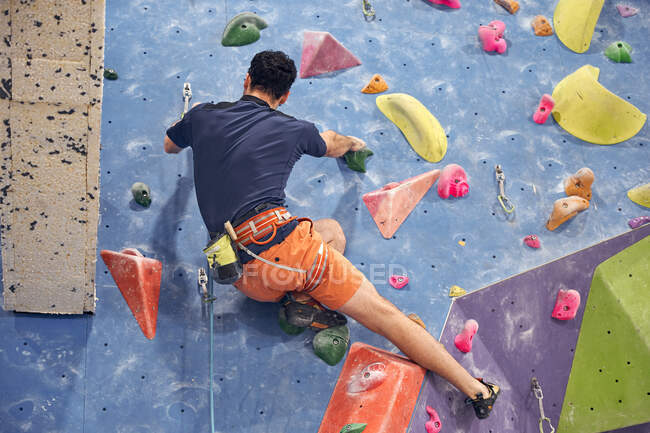Back view of anonymous male mountaineer in harness climbing artificial wall in bouldering center — Stock Photo
