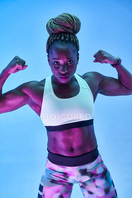 Athlète féminine afro-américaine musclée au corps moite montrant des biceps sur fond bleu — Photo de stock